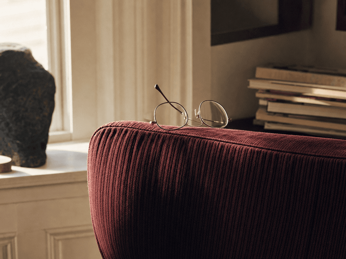Une paire de lunettes à monture fine posée sur un dossier de fauteuil en velours côtelé bordeaux, dans la lumière oblique d'une fenêtre d'intérieur.